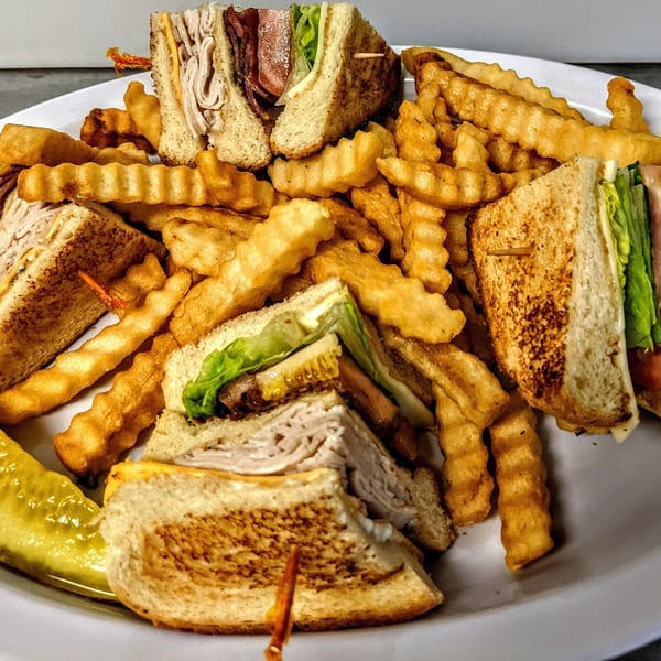 A Waffle Shoppe club sandwich, cut into quarters, served with crinkle-cut fries and a pickle spear.