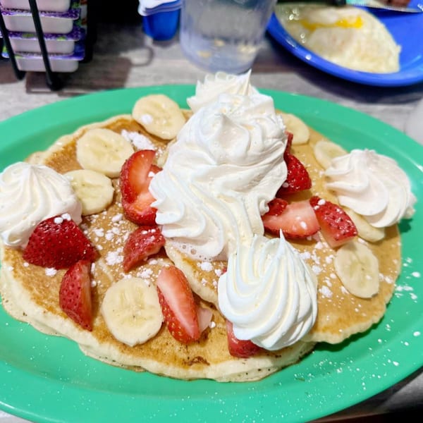Fluffy pancakes topped with whipped cream, sliced strawberries, bananas, and powdered sugar on a green plate.