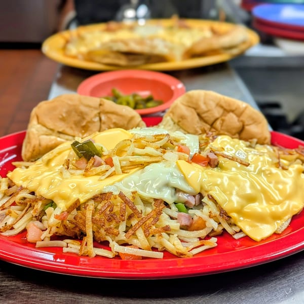 A Waffle Shoppe breakfast platter with hash browns, melted cheese, ham, and two buns on a red plate.